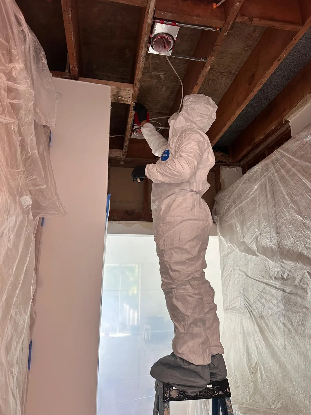Mold & Allergy technician in a full Tyvek suit performing ceiling demolition inside a sealed plastic containment area.